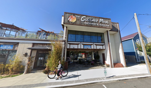 A woman rides a bike past The Cultured Pearl restaurant, featuring a modern facade and outdoor seating area.