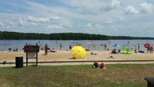 A sandy beach with people swimming and sunbathing, colorful umbrellas, and a calm lake under a partly cloudy sky.