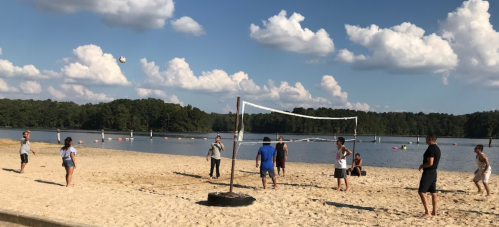 A group of people playing beach volleyball on a sandy shore with a lake and clouds in the background.