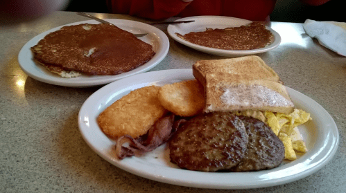 A breakfast plate with scrambled eggs, sausage, bacon, hash browns, toast, and two large pancakes in the background.