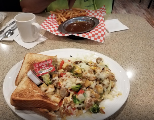 A plate of cheesy vegetable hash with toast and a side of fries and gravy on a table. Coffee cup in the background.
