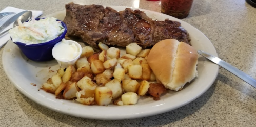 A plate with a grilled steak, seasoned potatoes, a roll, and a side of coleslaw.