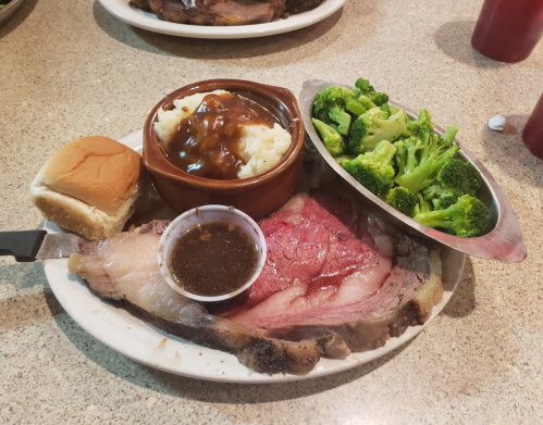 A plate with prime rib, mashed potatoes with gravy, steamed broccoli, and a dinner roll on a table.