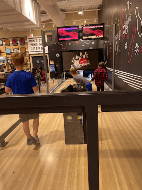 Children playing bowling at an indoor bowling alley, with screens displaying scores in the background.