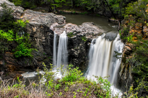 A scenic view of a waterfall cascading over rocks into a calm river, surrounded by lush greenery.