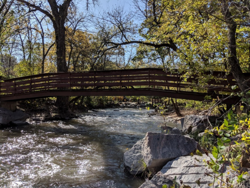 A wooden bridge spans a flowing river, surrounded by trees and rocks under a clear blue sky.