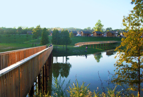A wooden walkway extends over a calm lake, surrounded by greenery and cabins in the background under a clear sky.
