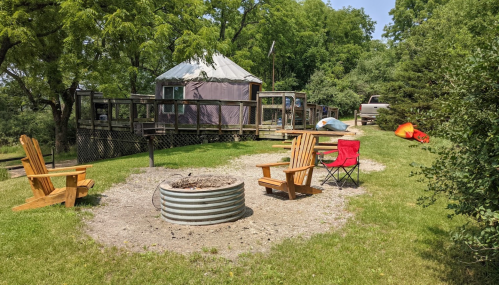 A cozy campsite with a fire pit, wooden chairs, and a yurt in the background surrounded by greenery.