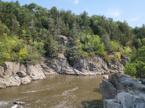 A serene river flows past rocky cliffs, surrounded by lush green trees under a clear blue sky.