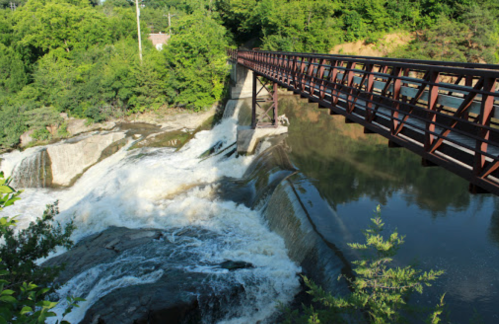 A metal bridge spans a waterfall, surrounded by lush greenery and reflecting in the water below.