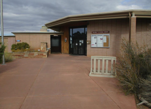 Entrance of a visitor center with a wooden door, bulletin board, and desert landscaping under a cloudy sky.