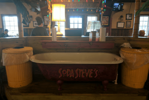 A vintage bathtub labeled "Soda Steve's" sits between two yellow trash cans in a casual dining setting.