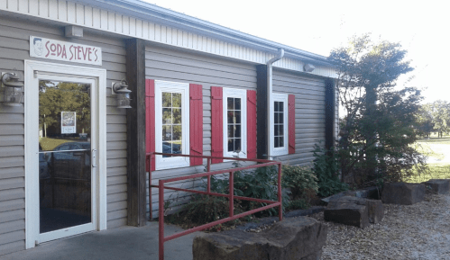Exterior of Spa Steve's, featuring a sign, red shutters, and a ramp leading to the entrance. Lush greenery surrounds the building.