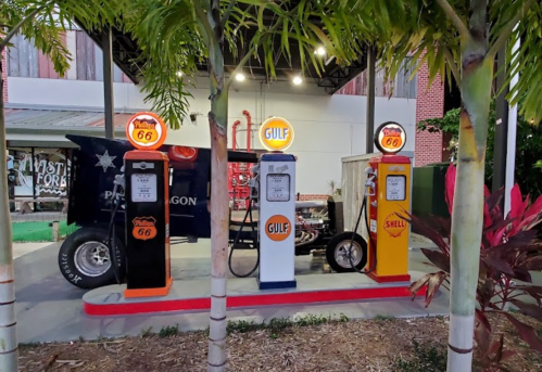 Vintage gas station with retro pumps from Gulf and Shell, alongside a classic car, surrounded by tropical plants.