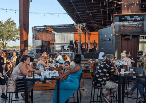 A lively outdoor venue with people dining and enjoying live music on stage, surrounded by trees and string lights.
