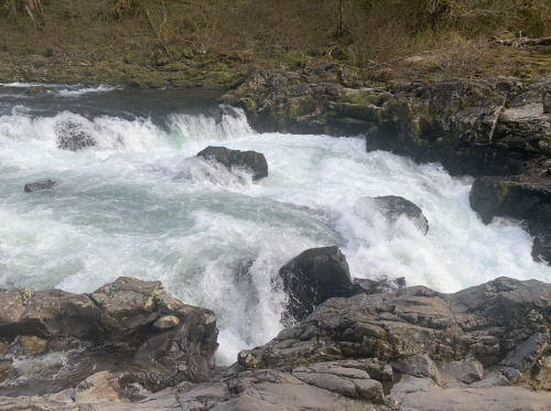 Rushing water flows over rocky terrain in a natural setting, surrounded by greenery and rugged landscape.