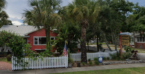 A charming restaurant with a red exterior, surrounded by palm trees and a white picket fence, on a sunny street.