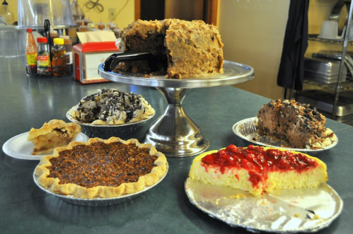 A variety of desserts on a table, including cakes, pies, and a cheesecake, displayed on plates and a cake stand.