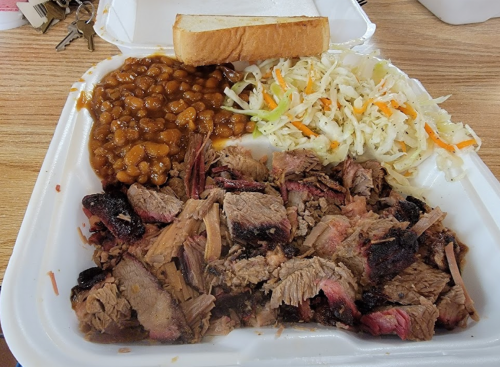 A takeout container with sliced smoked brisket, baked beans, coleslaw, and a slice of bread.