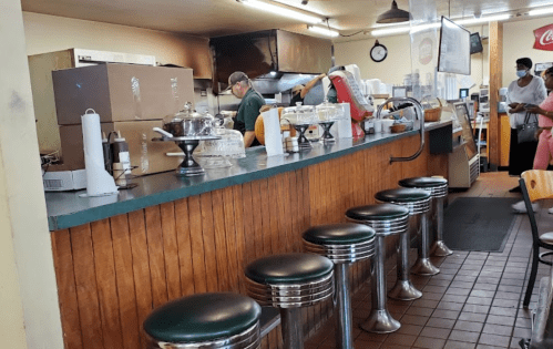 A diner counter with green stools, glass displays, and staff preparing food in a cozy, casual setting.
