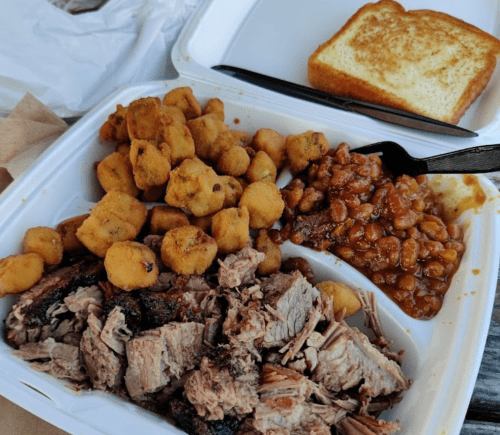 A takeout container with sliced brisket, fried okra, baked beans, and a slice of toasted bread.