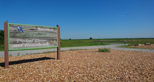 Sign welcoming visitors to Pintail Drive and Boardwalk at a national wildlife refuge, with a clear blue sky and green fields.