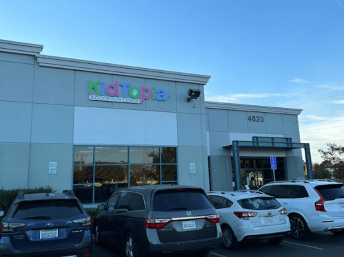 Exterior of KidTopia Indoor Playcenter, featuring colorful signage and parked cars in front.
