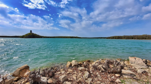 A serene lake with turquoise water, rocky shore, and a distant green hill under a partly cloudy sky.