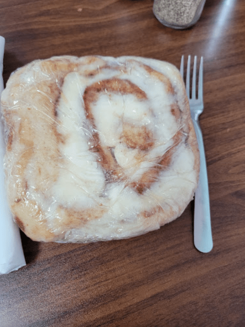 A cinnamon roll wrapped in plastic, placed on a wooden table next to a fork and a salt shaker.