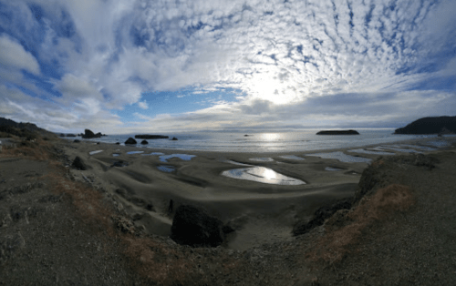 A panoramic view of a sandy beach with tidal pools, under a cloudy sky and distant islands on the horizon.