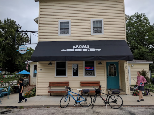 Aroma Pie Shoppe storefront with a black awning, two bicycles parked outside, and people walking by.