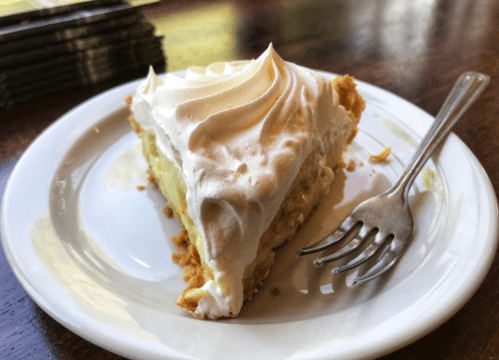 A slice of banana cream pie topped with whipped cream on a white plate, with a fork beside it.
