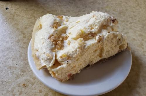 A slice of creamy dessert topped with crushed nuts on a white plate, resting on a textured table surface.