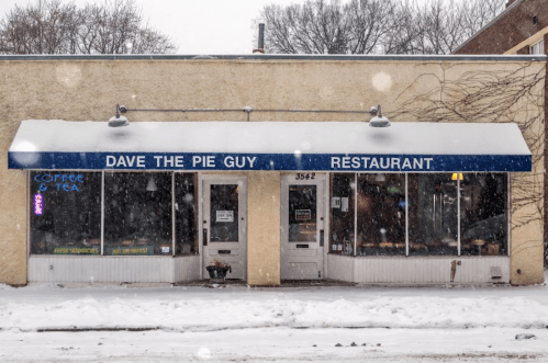 A snowy scene of "Dave the Pie Guy" restaurant with a blue awning and large windows, showcasing a cozy atmosphere.