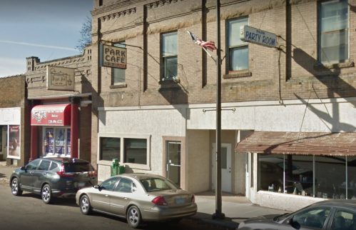 Street view of a small town with a café, party room, and parked cars under a clear blue sky.