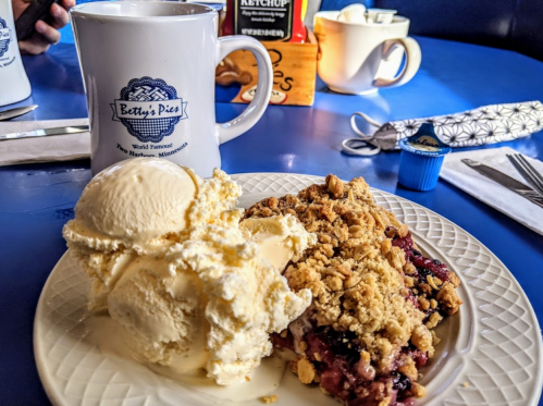 A plate with warm berry crumble topped with vanilla ice cream, alongside a coffee mug on a blue table.