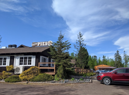 A rustic building with a sign reading "Fresh Pies," surrounded by trees and parked cars under a blue sky.