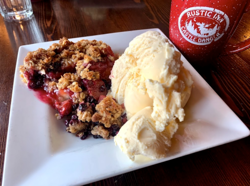 A slice of berry crumble dessert served with a scoop of vanilla ice cream on a white plate, next to a red mug.