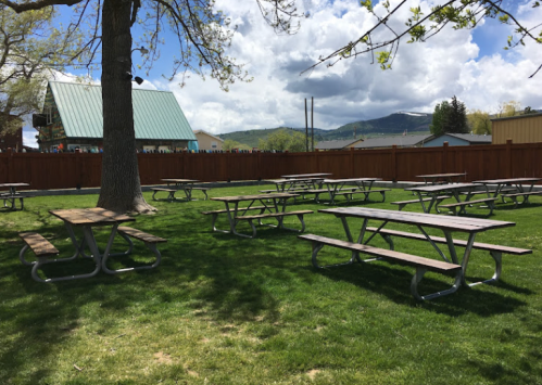 A grassy area with several picnic tables under a tree, surrounded by a wooden fence and mountains in the background.