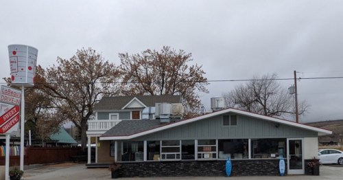 A small, single-story restaurant with a gray exterior and a sign, surrounded by trees and a cloudy sky.