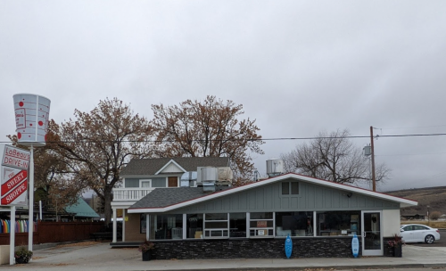 A small restaurant with a gray exterior and a sign, surrounded by trees and a cloudy sky.