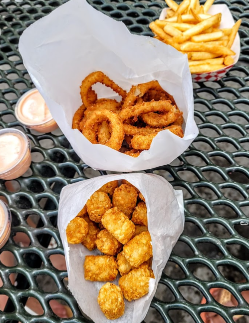 A close-up of two paper containers: one with crispy onion rings and the other with golden tater tots, alongside fries.