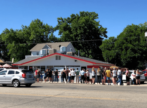 A long line of people waiting outside a restaurant on a sunny day, with cars parked nearby and trees in the background.
