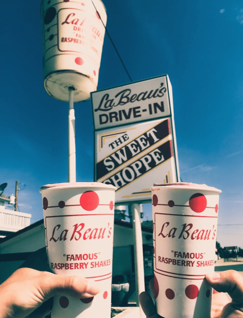 Two hands hold large cups of raspberry shakes in front of a drive-in sign that reads "La Beau's Sweet Shoppe."