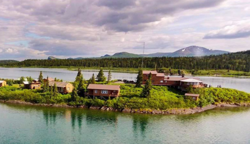 Aerial view of a small island with rustic cabins surrounded by a calm lake and lush greenery under a cloudy sky.