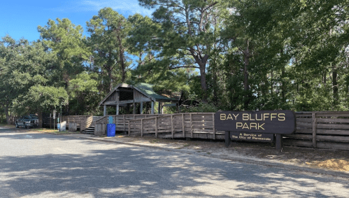 Sign for Bay Bluffs Park, surrounded by trees, with a wooden structure and parking area visible in the background.