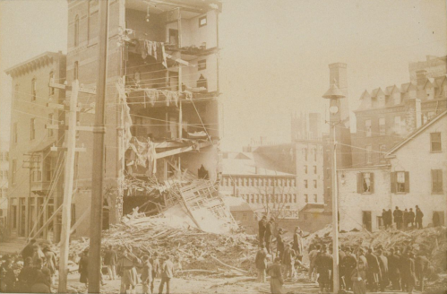Historic photo of a collapsed building, with debris scattered and onlookers gathered nearby.