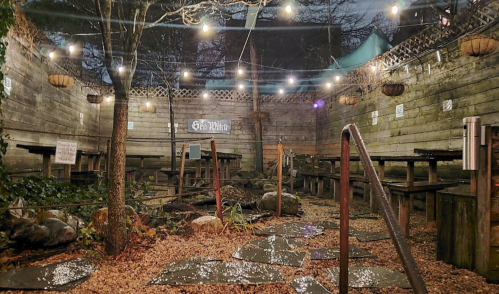 A cozy outdoor seating area with string lights, wooden tables, and gravel paths, surrounded by trees at night.