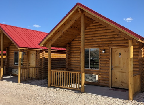 Two rustic log cabins with red roofs, featuring wooden porches and numbered doors, set against a clear blue sky.