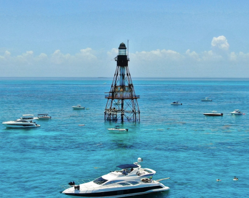 A lighthouse stands in clear blue waters, surrounded by boats under a bright sky with fluffy clouds.